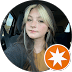 A young woman with long, light and dark blonde hair sits in a car, looking at the camera. She is wearing a yellow top and a dark outer layer. An orange and white star badge icon is in the lower right—perfect for a Denton Texas chiropractor ad.