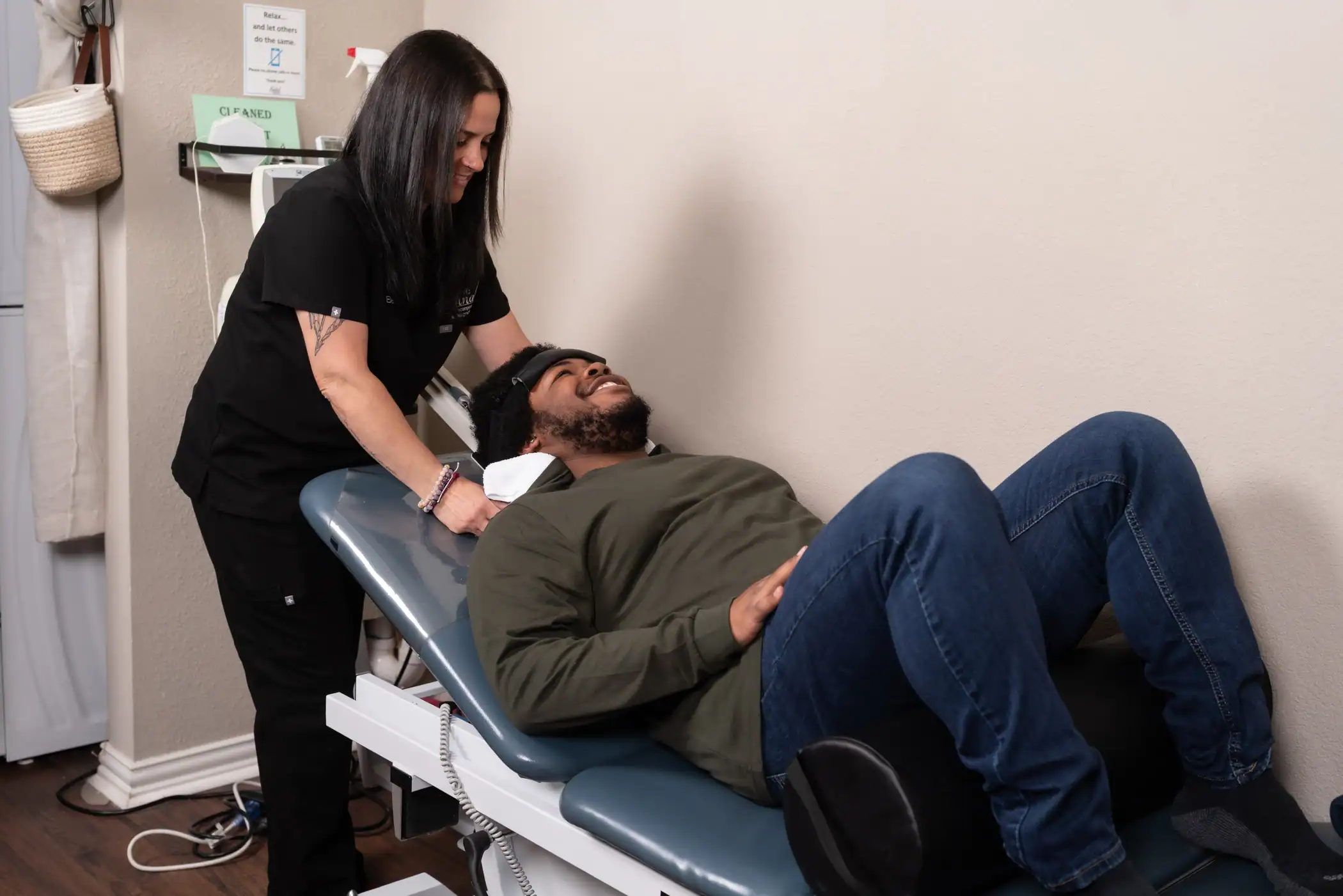 A woman in medical scrubs assists a man lying on an exam table, smiling as she adjusts a spinal decompression device on his neck in a clinic room.
