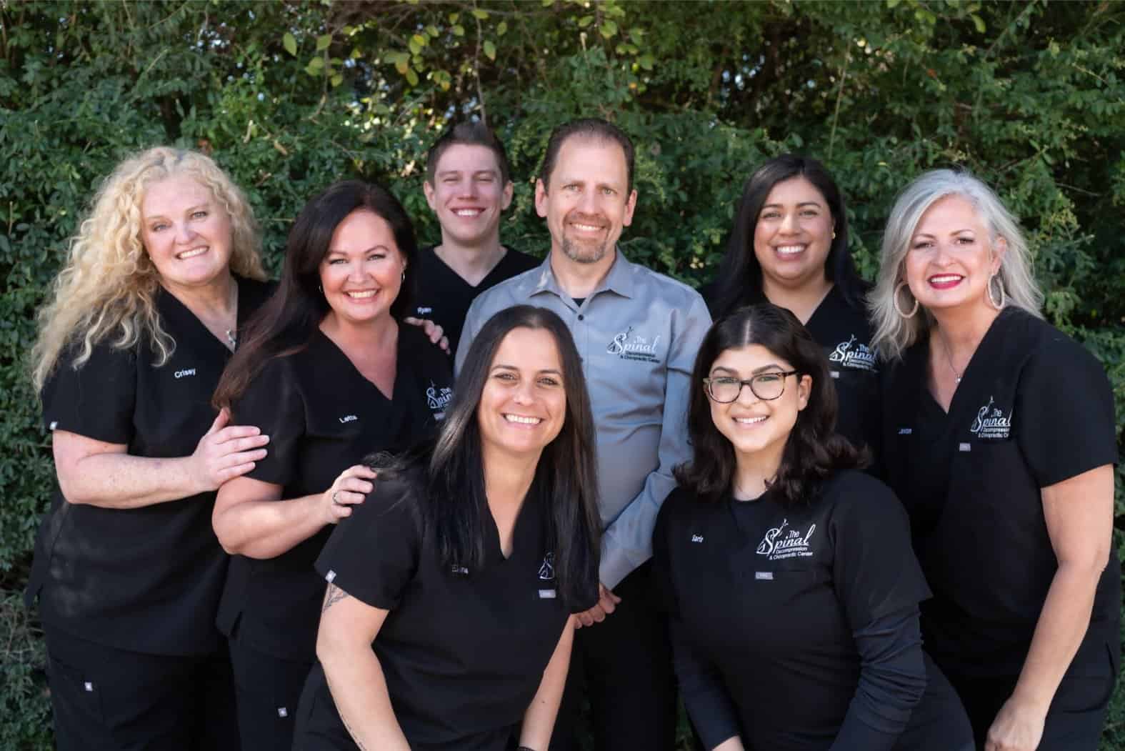 A group of eight smiling adults, most wearing black uniforms, pose together outdoors in front of green foliage. One man in a gray shirt stands in the center, surrounded by the Denton Texas chiropractor team specializing in spinal decompression.