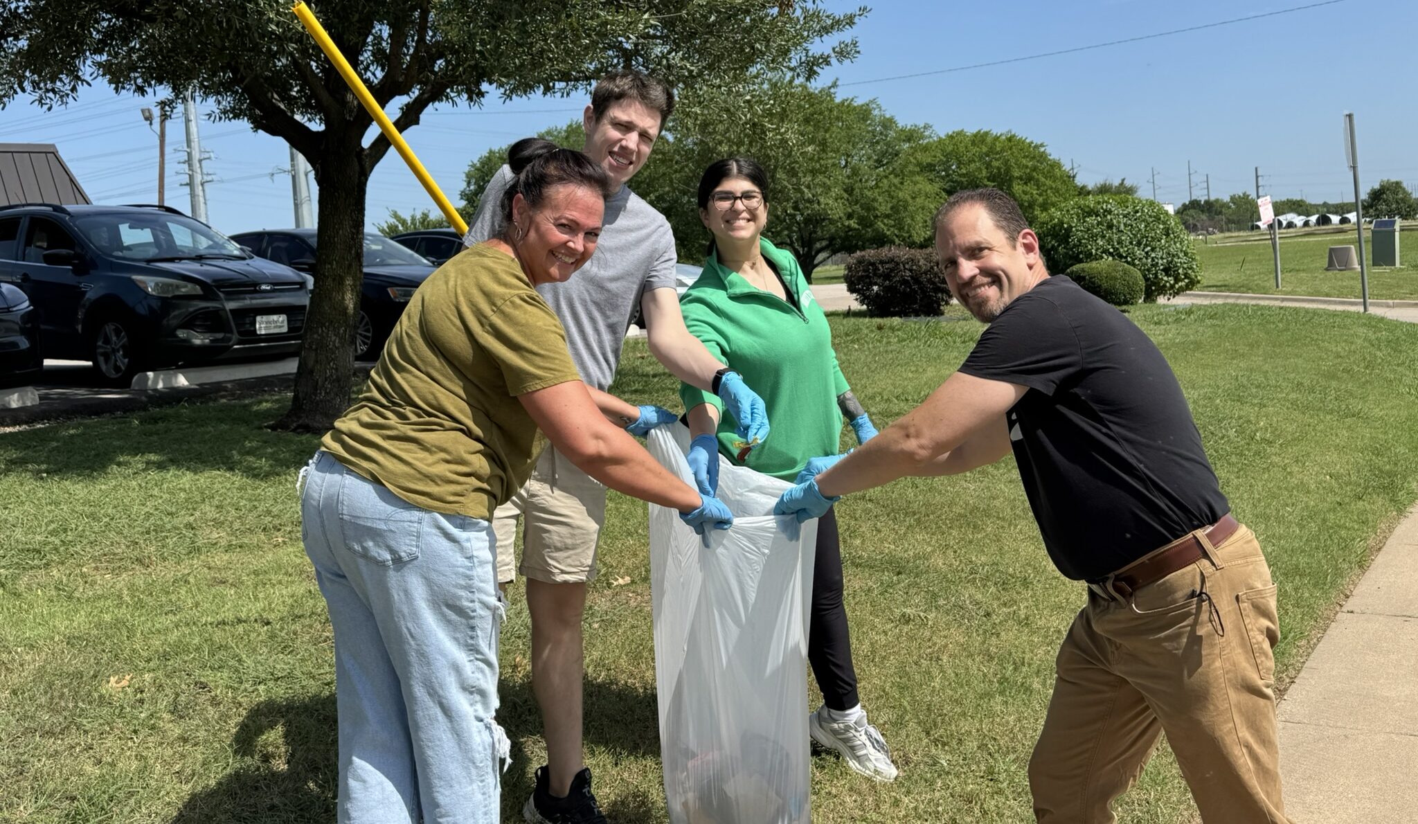 Four adults smiling and picking up litter together outdoors on a sunny day in Denton, Texas, placing trash into a large white bag. They are wearing gloves and standing on grass near parked cars and trees.