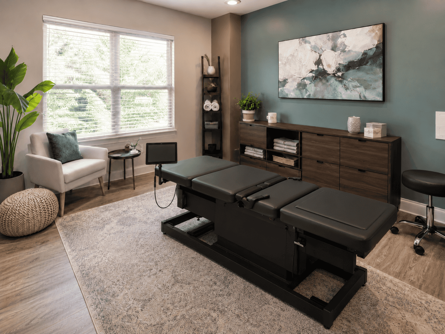 A modern chiropractor’s office in Denton with a black adjustment table, wooden cabinets, indoor plants, a white armchair, a pouf, and a large window with natural light. Abstract art hangs on a teal accent wall, creating an inviting space designed to guide patients through effective spinal decompression treatments.