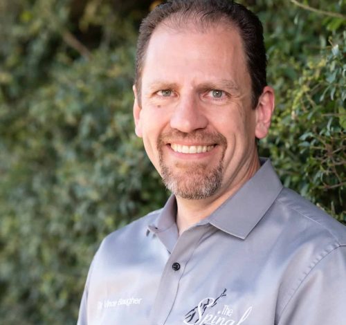 A man with short brown hair and a goatee, wearing a gray button-up shirt with embroidery, smiles while standing outdoors in front of green foliage.